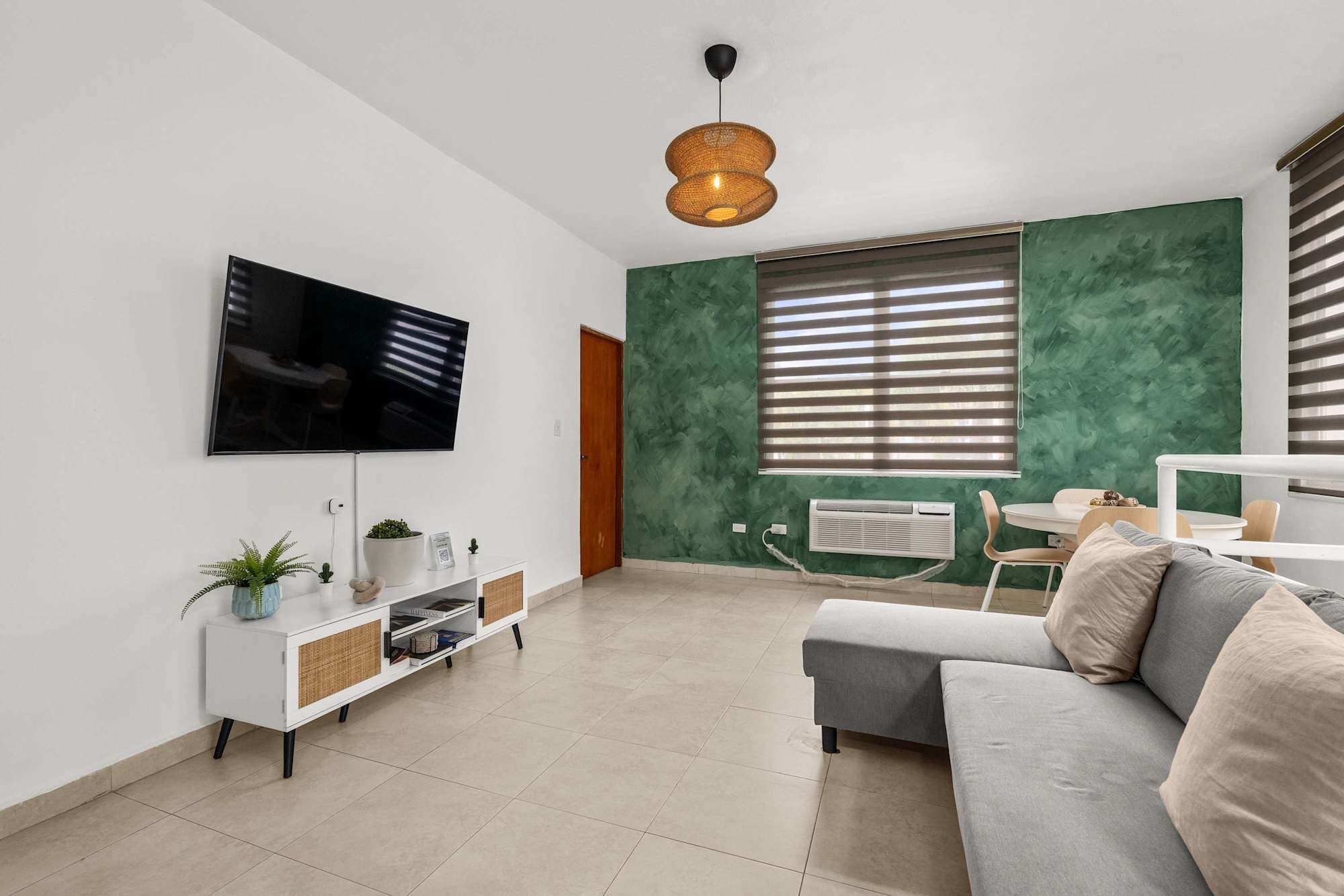 Sala con piso de baldosas claras. Televisor pantalla plana montado en pared blanca sobre consola baja con detalles de ratán. A la derecha, pared verde texturizada y parte de un sofá gris. Al fondo, comedor y ventana con persianas.