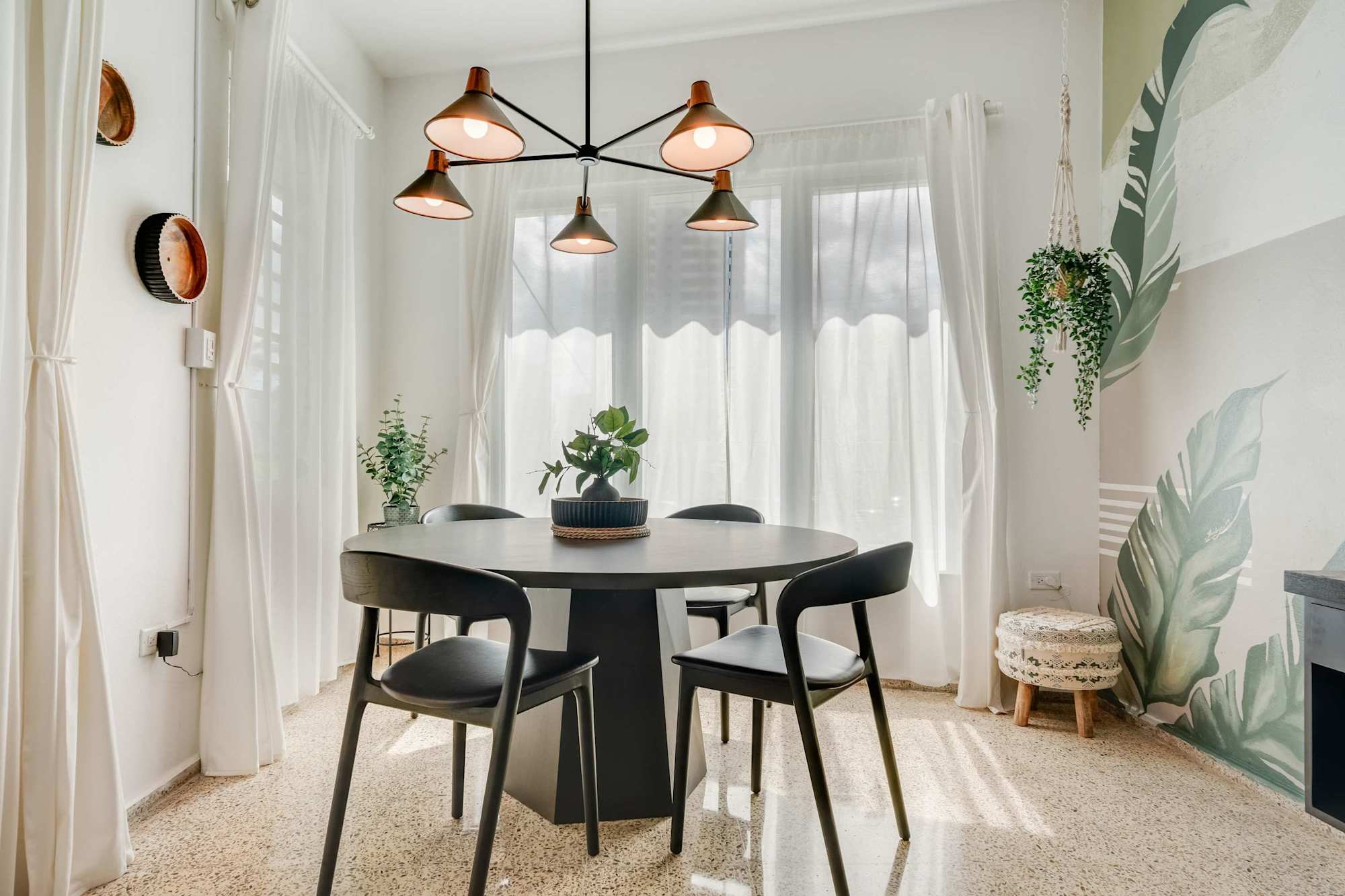 A dining area with a round black table and four matching chairs. Pendant lights hang above the table, and sheer white curtains cover the windows. Potted plants decorate the corners of the room.