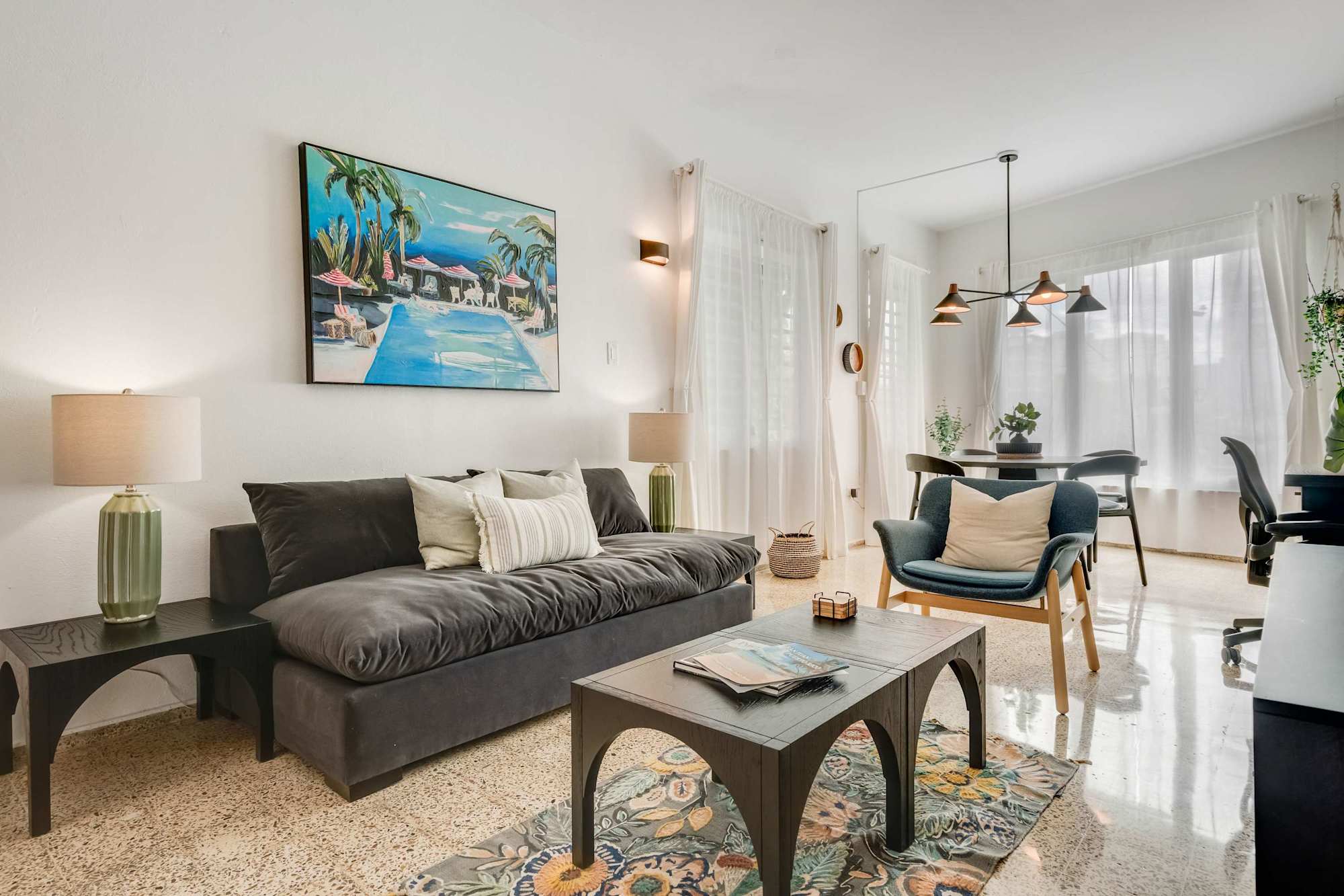 A living room with a dark sofa facing a wall-mounted TV. A small coffee table sits in front of the sofa, and open shelving with decorative items lines the wall. Neutral tones and natural light fill the space.