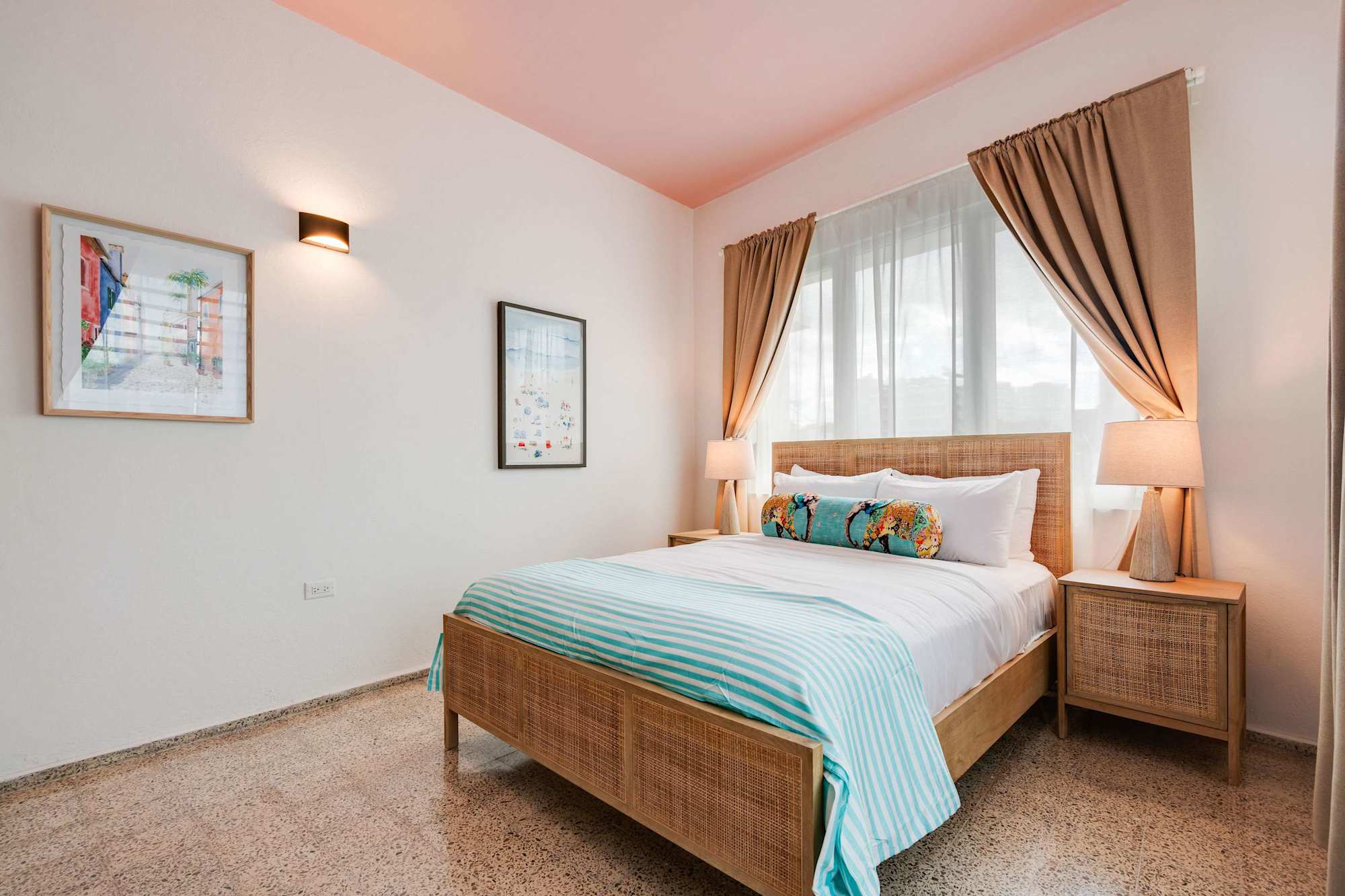 A bedroom with a queen-size bed dressed in white linens and a light blue throw. Two windows with beige curtains allow natural light. A wooden nightstand and soft lighting frame the bed.
