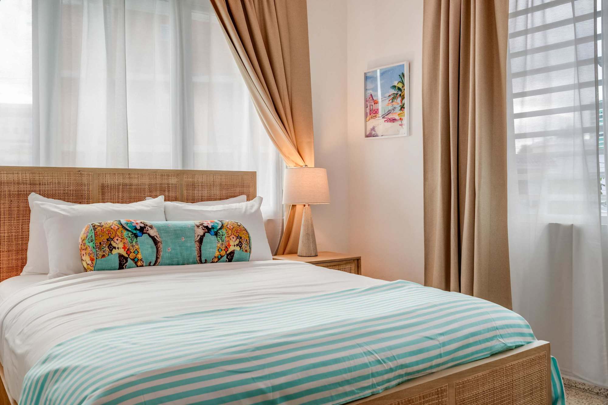 A bedroom with a queen-size bed dressed in white linens and a light blue throw. Two windows with beige curtains allow natural light. A wooden nightstand and soft lighting frame the bed.